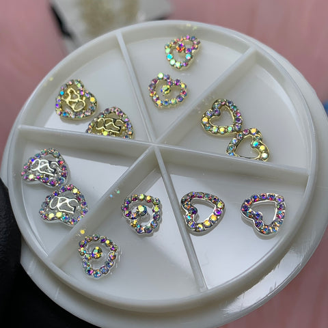 A round white container with a divided section holds Dynamic Nail Supply's 3D Gold Heart shape Charms, designed for Valentine's nail art. Each charm is adorned with colorful, iridescent rhinestones, ideal for adding intricate and sparkling designs to your nails. The charms are arranged neatly in the compartments.