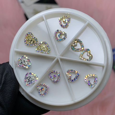 A round container with six compartments from Dynamic Nail Supply holds 3D Gold Heart shape Charms, ideal for Valentine's nail art designs. Each charm is embellished with small, glittering multicolored rhinestones. A gloved hand displays the container against a soft pink surface in the background.