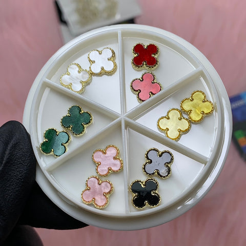 A hand in black gloves holds a circular white container divided into sections, each featuring flower-shaped charms from the Luxury 3D V4n-Cleef Charms set by Dynamic Nail Supply. The paired charms display vibrant colors such as red, yellow, and gold against a soft pink background.