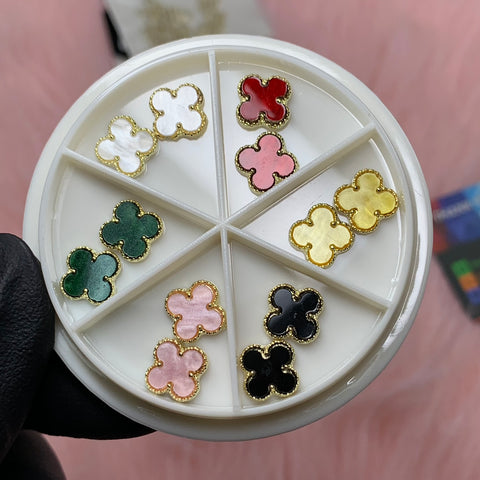 A round white container features nine clover-shaped Luxury 3D V4n-Cleef Charms with gold borders, available in a variety of colors including white, red, pink, yellow, green, and black. A gloved hand showcases this impressive Dynamic Nail Supply set against a pink textured background.