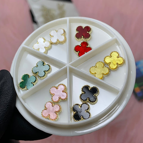 A close-up image features a hand holding a circular tray from the Luxury 3D V4n-Cleef Charms set by Dynamic Nail Supply. The white tray, divided into six compartments, displays clover-shaped earrings in various shades—white, red, green, pink, black, and yellow—against a pink fuzzy background.