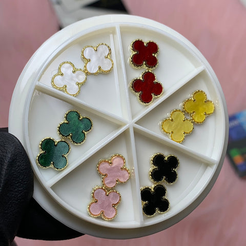 The Luxury 3D V4n-Cleef Charms set from Dynamic Nail Supply is a circular container with six compartments that houses pairs of clover-shaped nail art in white, red, green, yellow, pink, and black. Each charm has a gold outline and exudes luxury. The set is held by a hand wearing a black glove.