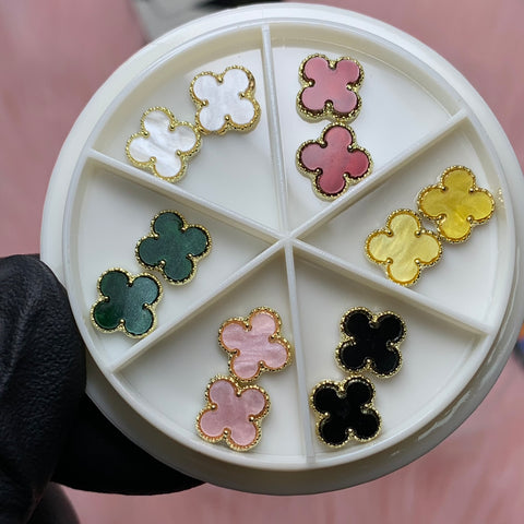 A white circular tray from the Luxury 3D V4n-Cleef Charms set by Dynamic Nail Supply displays six pairs of flower-shaped stud earrings in a range of colors including white, pink, yellow, green, light pink, and black. Each earring is accented with a gold-colored edge. The tray is shown being held by someone wearing a black glove against a soft pink background.