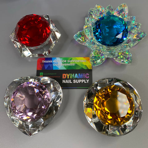 Four intricately designed, large lead crystal gems in red, blue, purple, and yellow are elegantly displayed around a Dynamic Nail Supply business card. The blue crystal is mounted on a lotus-shaped base. The business card features a rainbow background with a thank you message on a light gray surface.