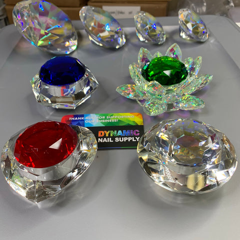 A stunning arrangement of large, multifaceted lead crystal stones in hexagon, round, heart, and lotus shapes features prominently. Each stone showcases a central gem in blue, red, or green hues encircled by clear crystals arranged like petals. These Crystal Dappen Dishes are elegantly displayed on a table accompanied by a small card that reads Dynamic Nail Supply. The entire setup brilliantly reflects light.