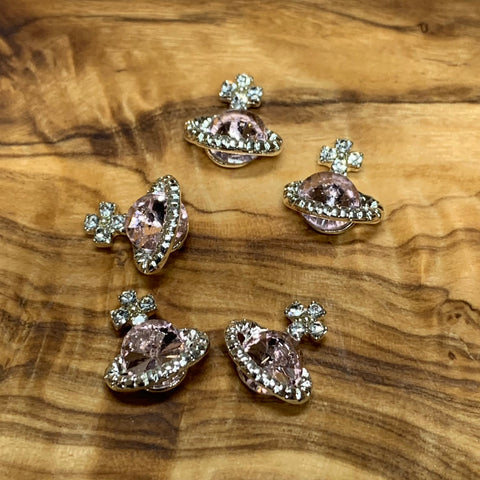 Five gold-tone brooches, named "5 pcs Planet Charm for Nails art designer" by Dynamic Nail Supply, feature pink rhinestone centers shaped like planets with orbit rings, each adorned with small sparkling stones. These charms are displayed on a polished wooden surface and serve as a delightful inspiration for any nail art designer aiming to incorporate planet-themed charm into their creations.