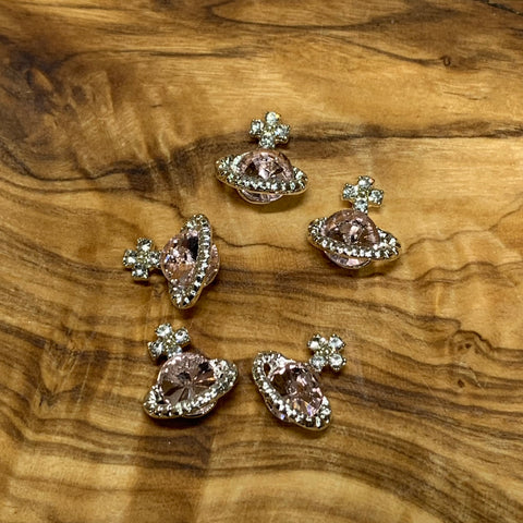 Five Planet Charms for nail art designers, from Dynamic Nail Supply, are displayed on a textured wooden surface. These pink rhinestone-engraved pins resemble Saturn with their ring designs and feature delicate floral motifs accented with clear gemstones for a touch of sparkle—ideal inspiration for any nail art designer.