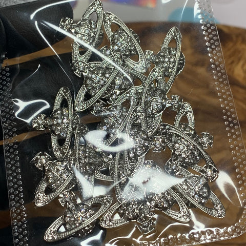 Close-up of a clear plastic bag containing the 20-piece set of Planet Charm for Nails from Dynamic Nail Supply. Each silver charm features a ring encircling a textured central globe adorned with small crystal rhinestones, resembling the planet Saturn. The background showcases a dark surface and part of the hand of an art designer holding the bag.