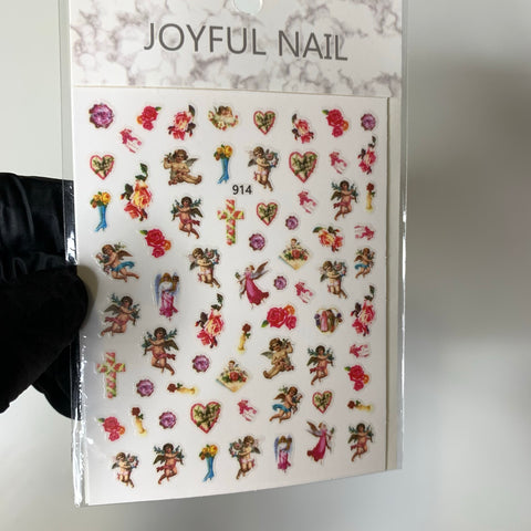 A hand in a black glove holds a pack of Dynamic Nail Supply's Religion Stickers, featuring angels and religious motifs such as flowers, hearts, and crosses. Each intricate design from the Angels - Mary Saint Sticker collection is neatly arranged on a white backing with the number 914 displayed.