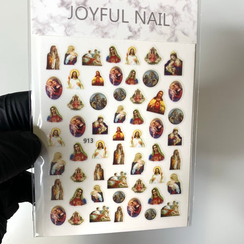 A sheet of Religion Stickers from Dynamic Nail Supply features vibrant images of Jesus, Mary Saint, and angels. The designs come in various colors, sizes, and shapes, including circular and oval formats. A gloved hand proudly holds the sheet labeled with "JOYFUL NAIL" at the top and numbered 913 at the bottom.