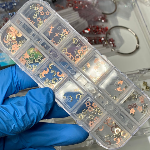 A gloved hand showcases a clear plastic organizer filled with Dynamic Nail Supply's Metal Sequins (SKU: 8894), featuring a variety of nail art decorations. Each compartment displays an assortment of metallic designs, including stars, moons, and hearts in alluring gold and rose gold tones. With 12 different styles available, these metal sequins add extra flair while additional containers subtly fade into the background.