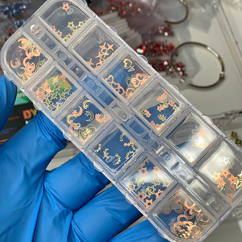 A gloved hand holds a transparent plastic container with multiple compartments, displaying 12 styles of metallic nail art decorations from Dynamic Nail Supply. Inside, the Metal Sequins (SKU: 8894) come in various shapes like stars, moons, and hearts. Red and gold decorations shimmer in the background.