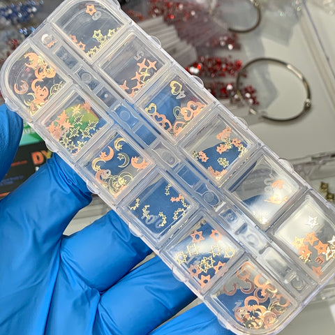 A gloved hand holds a clear plastic organizer containing 12 styles of metallic sequins for nail art, including designs like moons, stars, and hearts by Dynamic Nail Supply. In the background, blurred containers and a keyring are visible.