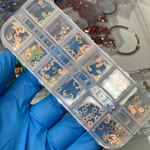 A person wearing a blue glove holds a transparent container, divided into small compartments, each containing 12 styles of gold and orange celestial-themed nail art pieces shaped like stars and moons. The background displays additional containers of Dynamic Nail Supply's Metal Sequins (SKU: 8894) for nail art designs.