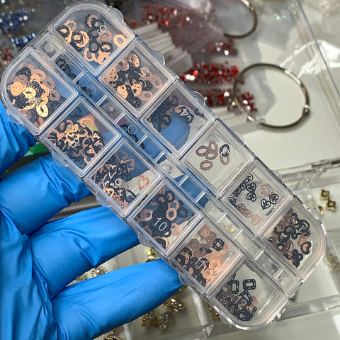 A hand adorned in a blue glove presents a clear plastic container featuring 12 assorted styles of small black and copper metallic charms from Dynamic Nail Supply's Metal Sequins (SKU: 8907), perfect for jewelry-making or nail art designs. Each section is filled with delightful flowers, hearts, and numbers, while additional supplies like beads and rings linger creatively in the background.