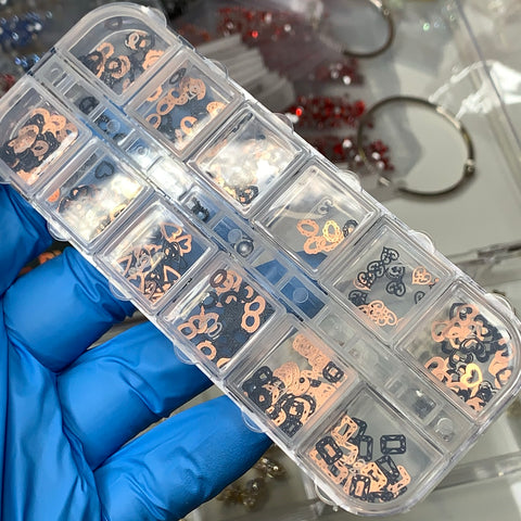 A hand in a blue glove holds a clear plastic container with compartments, each filled with Dynamic Nail Supply's Metal Sequins (SKU: 8907), featuring small metallic numbers and symbols in gold and black. Among the blurred metallic jewelry items in the background, these pieces shine as exquisite inspirations for your next nail art design.