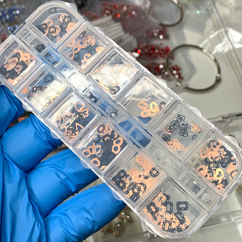 A gloved hand holds a clear plastic organizer filled with small nail art decorations in assorted shapes and colors, including pink and black. Among them are the "Metal Sequins" by Dynamic Nail Supply (SKU: 8907), offering 12 different styles. The background features blurred containers and trinkets, with hints of red and silver items visible.