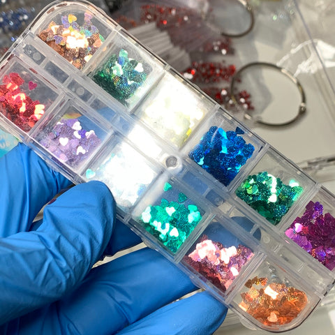 A person wearing a blue glove holds a plastic container featuring twelve compartments filled with Heart Shape Holographic Glitters / Sequins by Dynamic Nail Supply (SKU: 8916). The sequins shimmer in hues of red, pink, yellow, blue, green, and orange. Perfect for creative nail art designs, metal rings and plastic bags can be seen in the background.