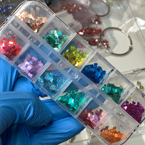 A person in blue gloves holds a clear plastic container with twelve compartments, each filled with colorful, reflective glitter star shapes ideal for nail art designs. These stars are complemented by Dynamic Nail Supply's Heart Shape Holographic Glitters (SKU: 8916), available in red, orange, yellow, green, blue, pink, and purple hues to add vibrant creativity.