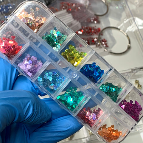 A close-up of a hand wearing a blue glove holds a segmented plastic container filled with Heart Shape Holographic Glitters from Dynamic Nail Supply, ideal for creative nail art designs. Each compartment features vibrant hues like pink, purple, green, blue, and red. Sequins are scattered in the background.