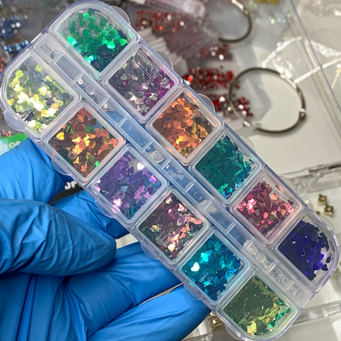 A hand in a blue glove holds a plastic organizer containing twelve compartments of Dynamic Nail Supply's Heart Shape Holographic Glitters / Sequins (SKU: 8924). Each compartment features vibrant shades such as pink, orange, green, blue, and purple. Jewelry-making supplies shine in the background, making them perfect for enhancing nail art designs.