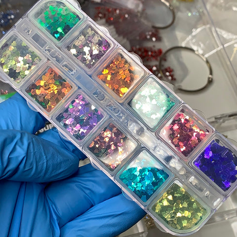 A gloved hand showcases a transparent plastic organizer featuring twelve compartments filled with Dynamic Nail Supply's Heart Shape Holographic Glitters (SKU: 8924). These bright sequins, ideal for nail art designs, offer a variety of shades including green, orange, purple, pink, and blue. In the background, metal hoops and red beads enhance the artistic charm.