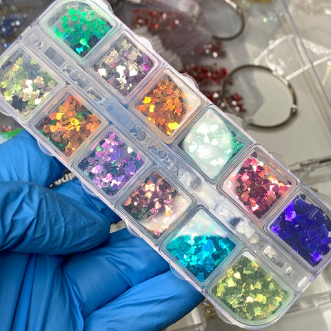 A gloved hand holds a clear plastic container, featuring twelve compartments, each filled with vibrant Heart Shape Holographic Glitters (SKU: 8924) from Dynamic Nail Supply. The sequins shimmer in shades of green, orange, pink, blue, and purple. Jewelry-making supplies and earring hoops are visible in the background, inspiring creative nail art designs.