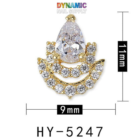 A luxurious piece from Dynamic Nail Supply's "Luxurious Jewelry for Nail Art Design" collection showcases a large, teardrop-shaped clear gem at the top, encased in a gold setting. Below it, two crescent rows of smaller clear gems add an extra touch of elegance. Measuring 11mm in height and 9mm in width, this exquisite charm is the epitome of high-end nail art. The text "HY-5247" is displayed beneath the charm.