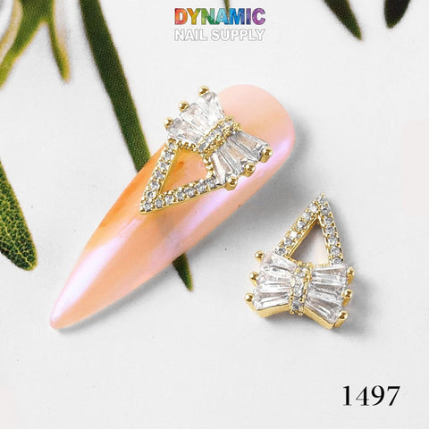 A close-up showcases a lavish nail art decoration featuring a prominent, gold-trimmed, clear gemstone bow adorning a pink, oval-shaped nail, capturing the essence of luxurious jewelry design. The identical decorative bow lies beside it on a white surface. The image is elegantly framed with green leaves and prominently displays the brand name Dynamic Nail Supply along with the number 1497. This is part of the Luxurious Jewelry for Nail Art Design - Part 2 collection.