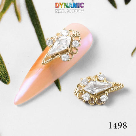 A decorative nail art piece from Dynamic Nail Supply, titled "Luxurious Jewelry for Nail Art Design - Part 2," features a peach-toned base adorned with intricate gold embellishments reminiscent of luxurious jewelry. The design includes a central diamond-shaped clear gem surrounded by smaller round gems, complemented by a matching decoration with the text 1498 displayed.