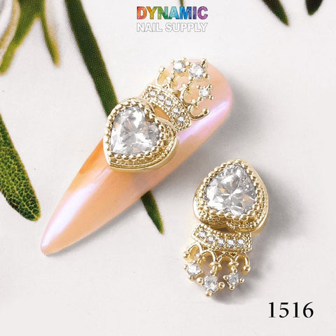 A close-up showcases two nail charms resembling gold and diamond, set against a pink nail background, radiating the essence of luxurious jewelry. Each charm is adorned with an elegant heart-shaped gem, complemented by a dangling design below. The image features "Dynamic Nail Supply" prominently at the top in multicolored text and displays "1516" at the bottom right. This design is part of Dynamic Nail Supply's "Luxurious Jewelry for Nail Art Design - Part 2.