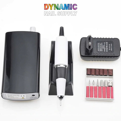 Displayed is the Manicure Nail Drill Machine - Electric File - Nail Tools by Dynamic Nail Supply. The set features a black and white handheld electric file on a stand, along with a black power adapter and a power box. Accompanying these is a compact case that holds six different drill bits and six sanding bands. The brand name "Dynamic Nail Supply" appears at the top in a colorful gradient effect.