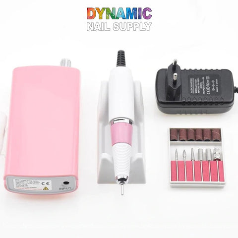 The Manicure Nail Drill Machine from Dynamic Nail Supply, perfect for manicures, is presented in pink and white. This set includes a pink power unit, a coordinating drill handpiece on a stand, a black power adapter, and interchangeable drill bits. The brand name Dynamic Nail Supply is prominently displayed above the image.