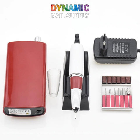 The image showcases the Manicure Nail Drill Machine - Electric File - Nail Tools from Dynamic Nail Supply. This vital set includes a red and white electric drill, a transparent cap, power adapter, and an assortment of nail tools neatly arranged in a plastic box. The brand's colorful logo is prominently displayed on the white surface backdrop.