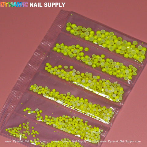 A pink surface showcases a plastic packet featuring the Round-shape Mixed-sizes Neon Rhinestone set, organized into five compartments each offering a variety of sizes. The brand name "Dynamic Nail Supply" is prominently displayed at the top in colorful text, with the website repeatedly mentioned at the bottom.