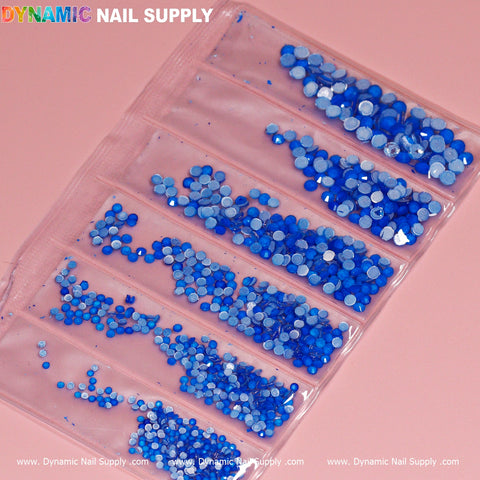 The Round-shape Mixed-sizes Neon Rhinestone set from Dynamic Nail Supply showcases assorted shades of blue nail art sequins, organized in six clear compartments against a pink background. Each section contains uniformly distributed neon rhinestones in various sizes, with hues spanning from light to dark blue.