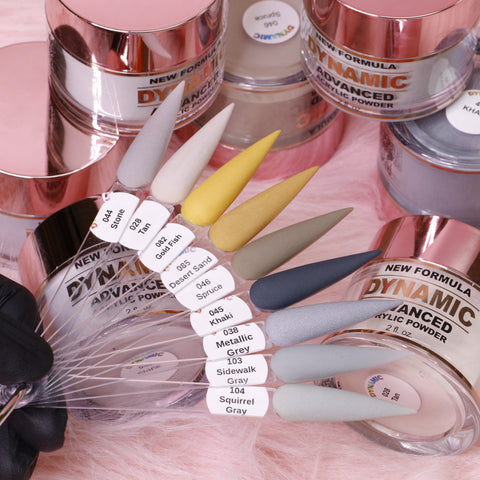 Close-up of the "Golden December" 2024 Acrylic Powder Collection from Dynamic Nail Supply, showcasing a variety of nail art colors labeled with names like Sidewalk Gray and Desert Sand. A hand displays clear swatches highlighting the range of hues from gray to blue tones. In the background, containers with rose gold lids ensure long-lasting nails.