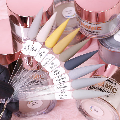 A fan displaying eight samples from the "Golden December" 2024 Acrylic Powder Collection highlights shades such as Stone, Desert Sand, Metallic Grey, and Squirrel Gray. In the background, containers of Dynamic Nail Supply's latest advanced acrylic powder promise long-lasting nails against a soft pink surface.