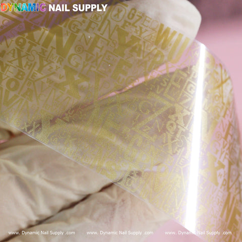 A gloved hand presents a semi-transparent sheet with embossed gold letters in various styles against a pink background. At the top left, the brand Dynamic Nail Supply is printed in vibrant colors, while the bottom border features their website, www.DynamicNailSupply.com, ideal for discovering the unique Nail Art Design Foils - Letters Style.