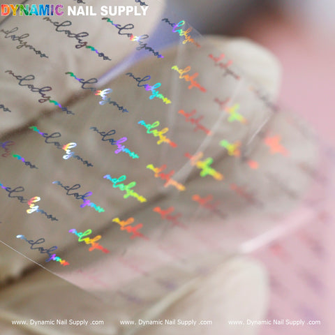 A close-up image displays a sheet from the Nail Art Design Foils - Letters Style collection, featuring holographic text that repeats "nail glam" in an elegant font. This vibrant nail art shimmers with blue, green, and red hues. Beige gloves hold the sheet, which prominently showcases the Dynamic Nail Supply brand at the top in a multicolored design.