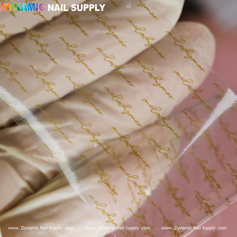 A gloved hand displays a transparent sheet adorned with the elegant repetition of the word "love" in gold cursive, highlighting intricate nail art. The blurred background accentuates the delicate design. In the top left corner, "Dynamic Nail Supply" is prominently featured in vibrant letters, and along the bottom edge are URLs related to Nail Art Design Foils - Letters Style.