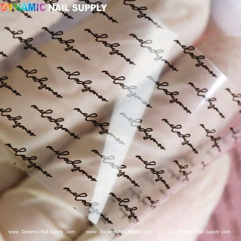 A transparent nail art film from Dynamic Nail Supply, part of their Nail Art Design Foils - Letters Style collection, showcases repeated cursive text reading "nailgasm" in black. The sheet is partially rolled on a soft-focus pink and white background. Branding for Dynamic Nail Supply is prominently displayed at the top and bottom edges in colorful text.