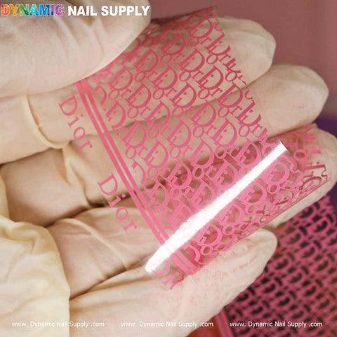 A close-up captures a gloved hand holding a transparent sheet from the Nail Art Design Foils - Luxury Style collection, featuring a pink pattern adorned with the word Dior. In the top left corner, Dynamic Nail Supply is prominently displayed. The blurred background reveals additional design foils.