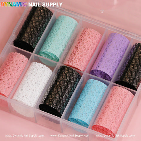 A plastic organizer containing nine compartments holds rolls of Nail Art Design Foils - Luxury Style from Dynamic Nail Supply. The colors available are mint green, coral pink, lavender, white, black, turquoise, and pale pink. These foils boast intricate lace-like patterns. Branding text from Dynamic Nail Supply adorns the top and bottom edges of the image.