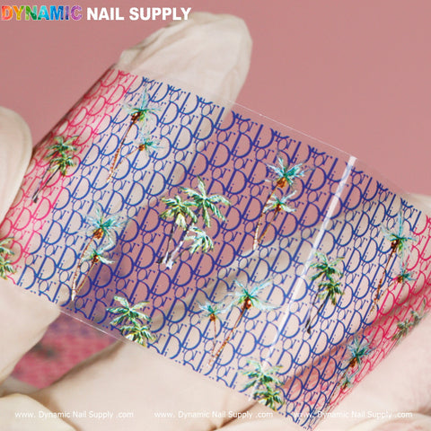 A gloved hand holds a clear film featuring a vibrant pattern with the letters DB in pink and blue, complemented by small palm trees—an ideal touch for nail art. The background is soft pink, and at the top of this luxury-style design foil image, you can see the brand name Dynamic Nail Supply.
