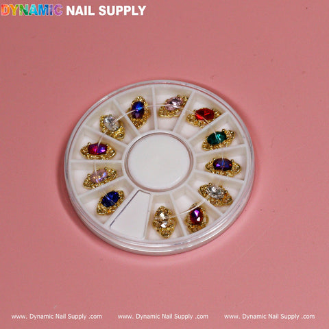 A round, white plastic wheel showcasing the 12 pcs Luxury Nail Charms boasts an array of luxury jewelry-style charms adorned with colorful gems such as purple, red, green, and clear stones. The wheel's segmented compartments contrast beautifully against a pink background. Above it all, Dynamic Nail Supply is elegantly written in vibrant letters.