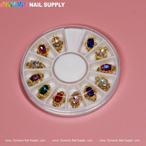 A round plastic container on a pink background displays the 12 pcs Luxury Nail Charms (Jewelry style) by Dynamic Nail Supply, featuring an assortment of gold-framed rhinestones. The vibrant stones in colors like red, blue, green, and clear are neatly organized in individual compartments. The brand name, Dynamic Nail Supply, is prominently featured at both the top and bottom edges.