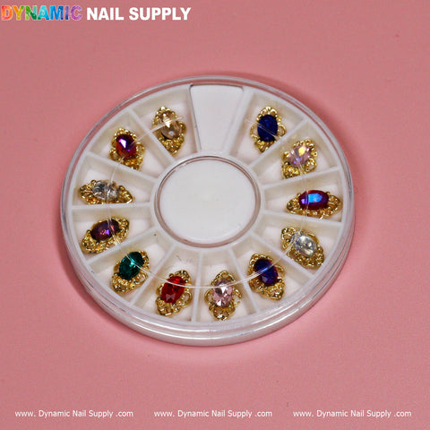 Displayed against a pink backdrop, the circular nail art wheel highlights 12 pieces of luxury nail charms in a jewelry style, featuring gold-edged gemstones in vibrant shades of red, blue, and green. Neatly organized in compartments, the packaging prominently features the Dynamic Nail Supply brand at both the top and bottom.