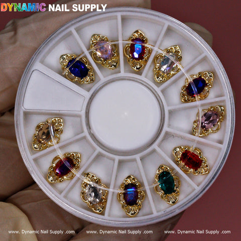 The 12 pcs Luxury Nail Charms by Dynamic Nail Supply come in a round container that beautifully displays an assortment of jewelry-style charms. Each piece is elegantly set in gold-toned metal, featuring colorful stones like red, blue, green, and pink. The container’s compartments highlight the intricate designs against a neutral background.