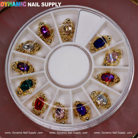 The Dynamic Nail Supply's 12 pcs Luxury Nail Charms provide a dazzling collection arranged in a round wheel with twelve compartments, each showcasing decorative jewelry-style charms in the shape of teardrops. Featuring vibrant colors such as red, pink, blue, and clear, each charm is encased in gold-tone frames with intricate details against a pink background.
